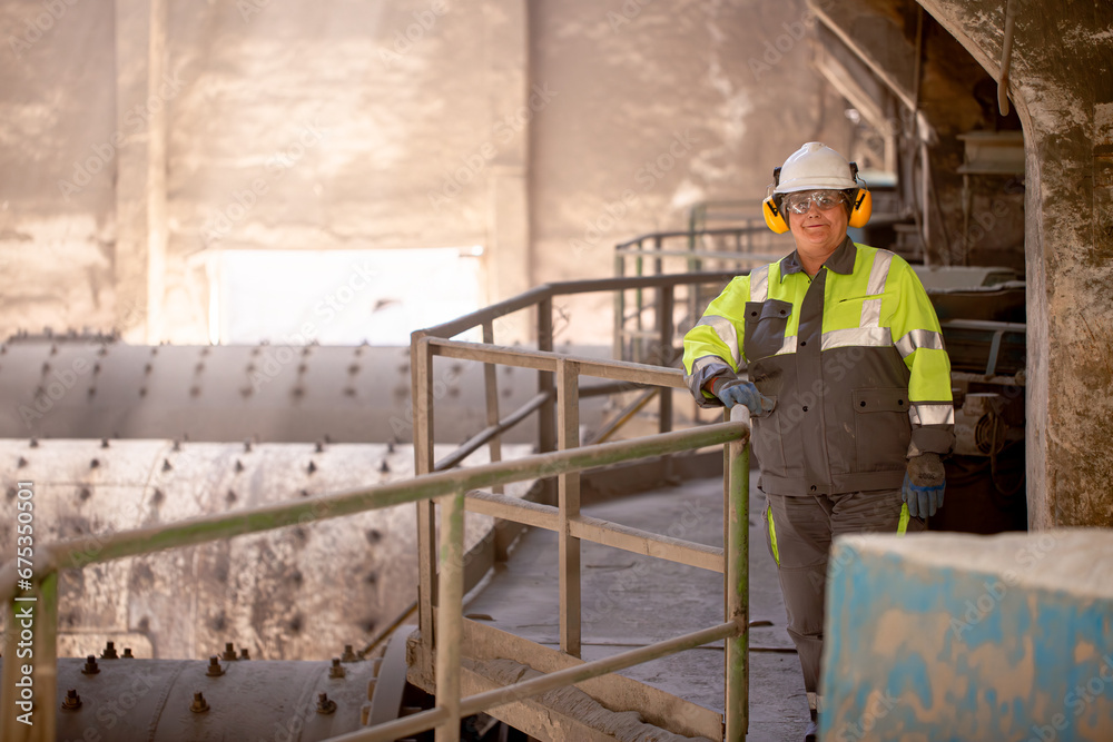 A worker in a helmet and equipment stands against the backdrop of a cement plant workshop. Work technology, cement production. People working in heavy industry. A worker in a helmet and equipment stands against the backdrop of a cement plant workshop. Work technology, cement production. People working in heavy industry.