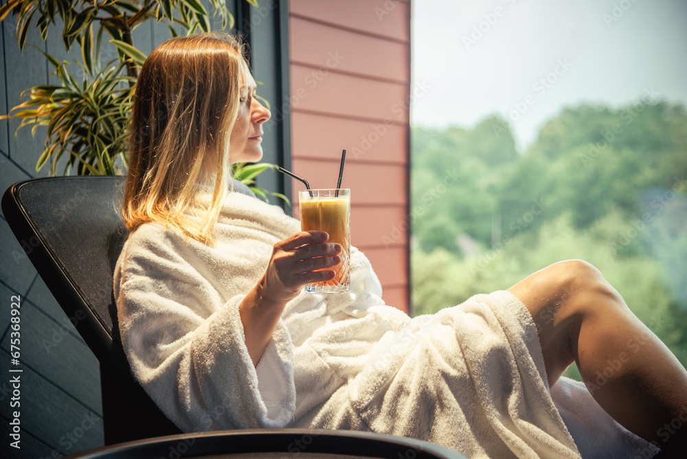 Woman in white bathrobe lying on sofa and relaxing with orange cocktail at SPA. - obrazy, fototapety, plakaty 