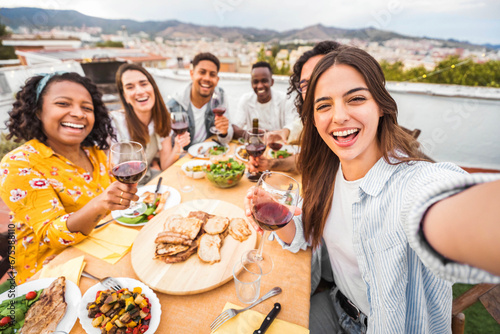 Happy friends having fun at rooftop dinner party - Group of young people taking selfie photo at outdoors dining table - Life style concept with guys and girls eating food and drinking wine together photo