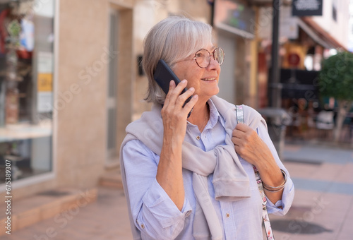 Smiling senior woman with white hair and eyeglasses walking in city street using cellphone. Beautiful elderly woman enjoys free time talking with smart phone © luciano
