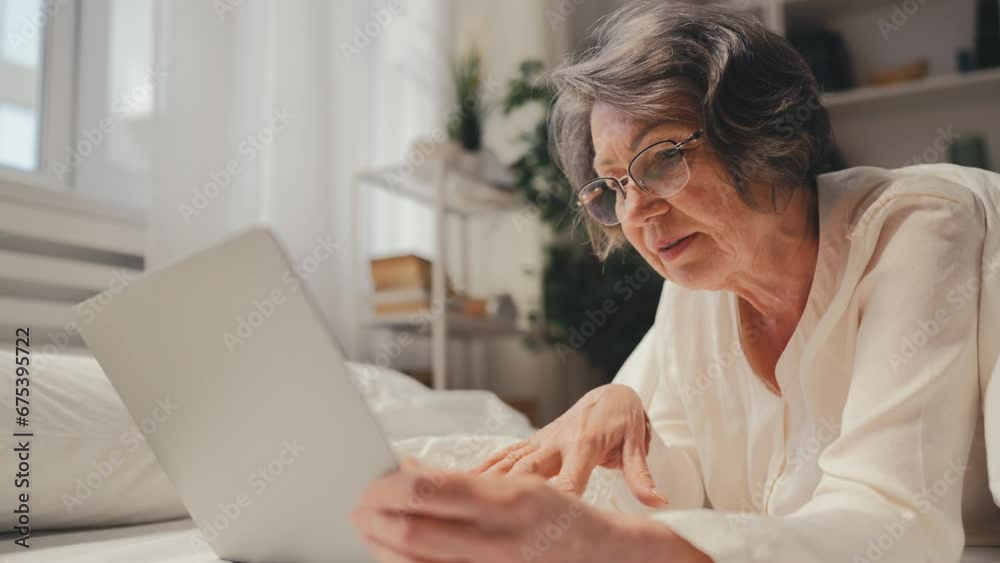 Woman in her 70s typing on laptop while lying in bed, freelancer lifestyle