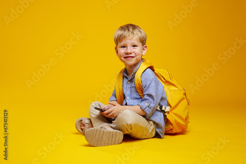 Quadro su tela Side view of excited pupil kid wearing blue shirt and yellow backpack, holding a phone in hands while sitting on studio floor with crossed legs, isolated over yellow background