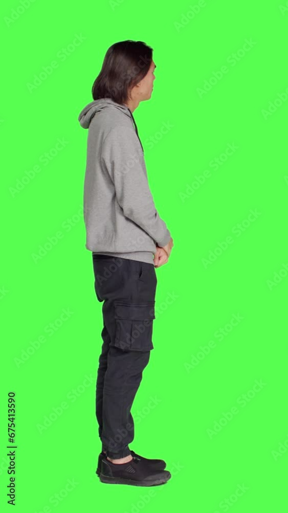 Profile in studio, young guy poses against greenscreen, looking at wristwatch and appearing distressed. Unhappy and irritated asian person is impatiently awaiting for stuff on camera. Profile in studio, young guy poses against greenscreen, looking at wristwatch and appearing distressed. Unhappy and irritated asian person is impatiently awaiting for stuff on camera.
