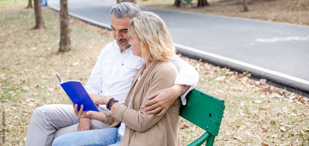 Caucasian senior couple reading book together on bench in the park.