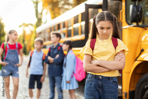 Unhappy young girl standing by herself next to yellow school bus