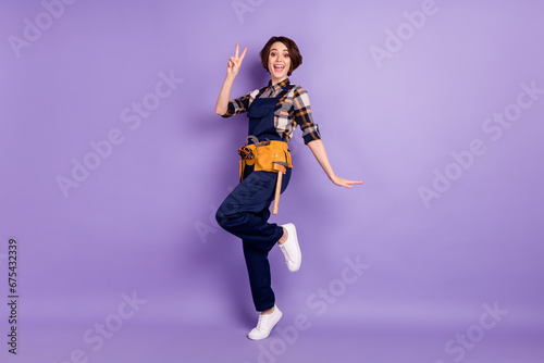 Full length body size photo of woman wearing overall showing v-sign gesture isolated on pastel purple color background