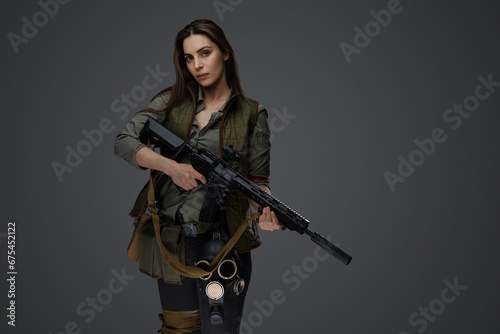 Middle Eastern-looking woman dressed in survivalist clothing posing with a rifle against a gray background, portraying strength and resilience