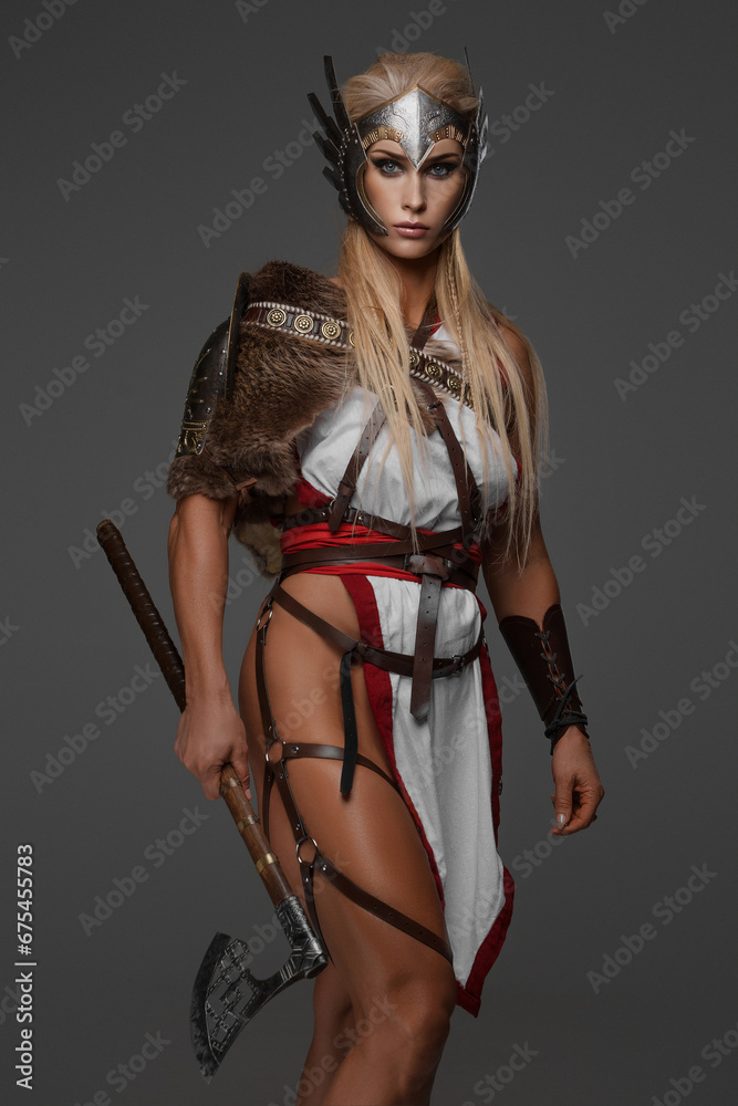 A muscular blonde Viking woman in fantasy armor poses holding two axes against a gray background A muscular blonde Viking woman in fantasy armor poses holding two axes against a gray background