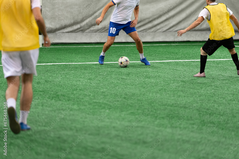 Five-a-side soccer players playing game in indoor stadium on artificial turf.