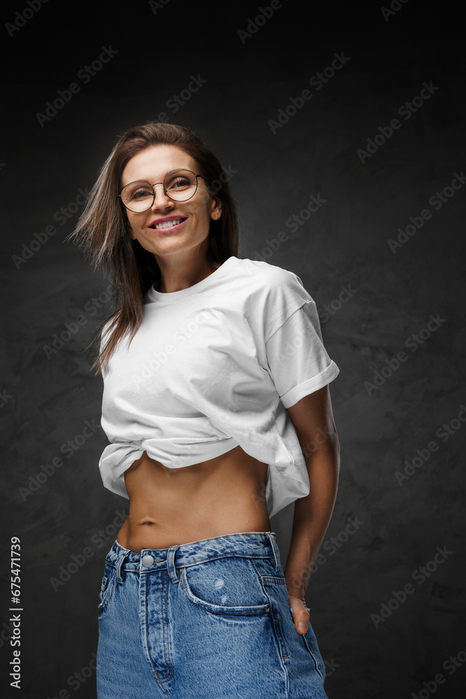 Gleaming woman in glasses and a white crop-top unveils her chiseled abs against a dark backdrop Gleaming woman in glasses and a white crop-top unveils her chiseled abs against a dark backdrop