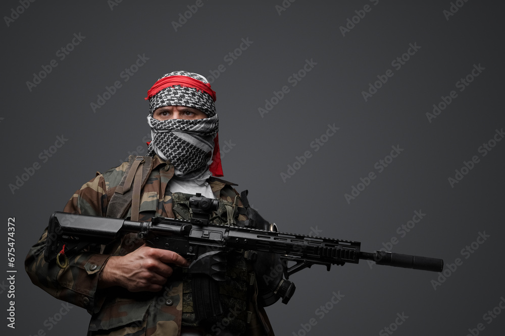 Fototapeta premium Militant from the Middle East, dressed in a white keffiyeh and camouflaged field attire, wielding an automatic rifle, set against a neutral gray backdrop