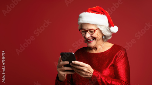 An elderly woman wearing a Santa hat and red dress is laughing joyfully while looking at her smartphone against a red background.