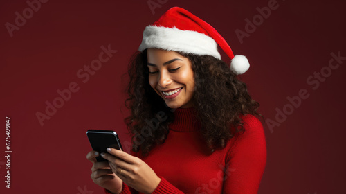 A young woman in a sweater and Santa hat is happily looking at her smartphone against a red background.