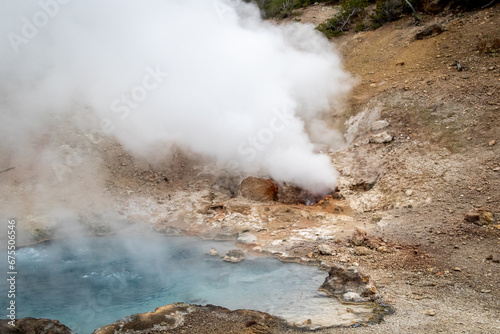 small geyser in yellowstone park national park, image shows a small gyser and a crystal clear blue heated pool of water, with sand and sulphur surrounding, taken october 2023