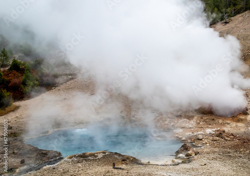 small geyser in yellowstone park national park, image shows a small gyser and a crystal clear blue heated pool of water, with sand and sulphur surrounding, taken october 2023