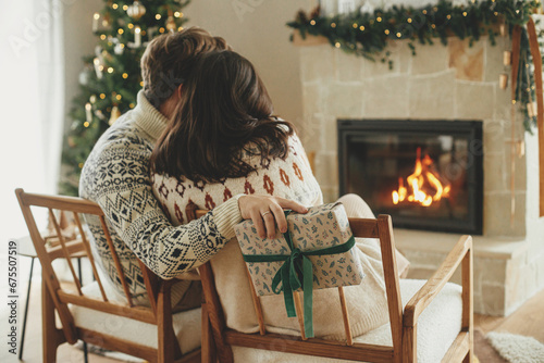 Happy young family in cozy sweaters exchanging stylish christmas gifts on background of fireplace with modern festive mantle and christmas tree with lights. Happy Holidays! photo
