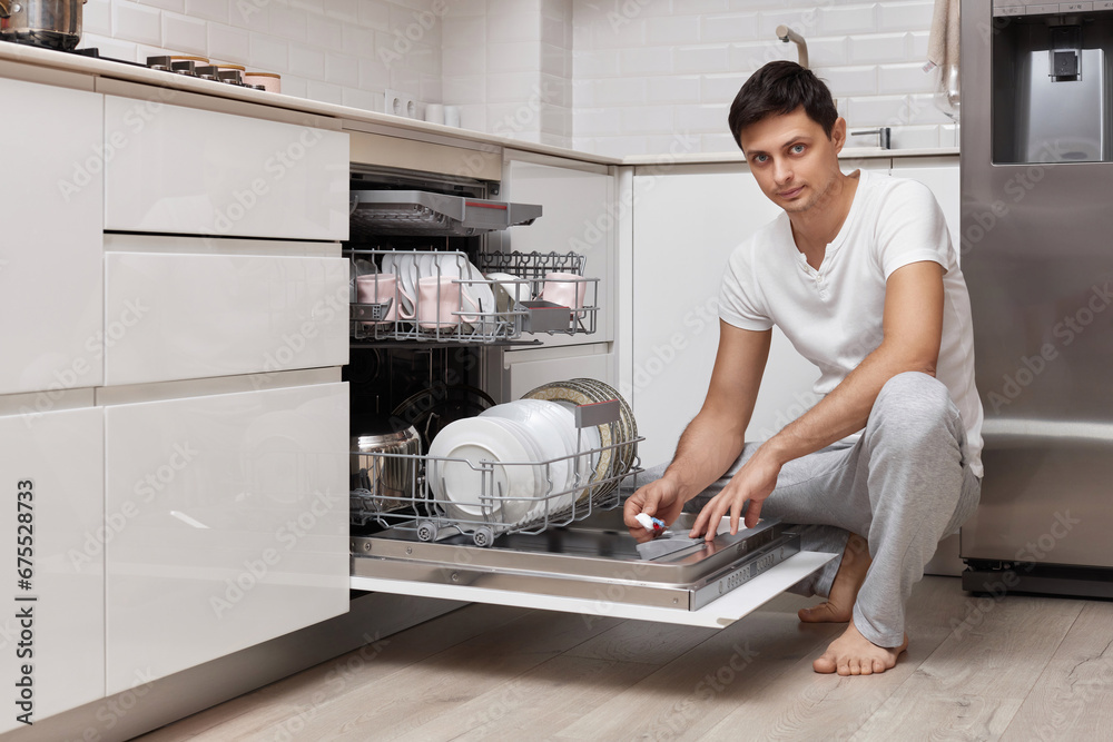 handsome man putting detergent tablet into dishwasher in kitchen