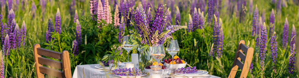 Elegant gorgeous wedding table decor or romantic dinner arrangement outdoors in blooming field. Purple lupine flowers, candles, fruits and wine, wooden furniture. Sunset, summer, golden hour banner Elegant gorgeous wedding table decor or romantic dinner arrangement outdoors in blooming field. Purple lupine flowers, candles, fruits and wine, wooden furniture. Sunset, summer, golden hour banner
