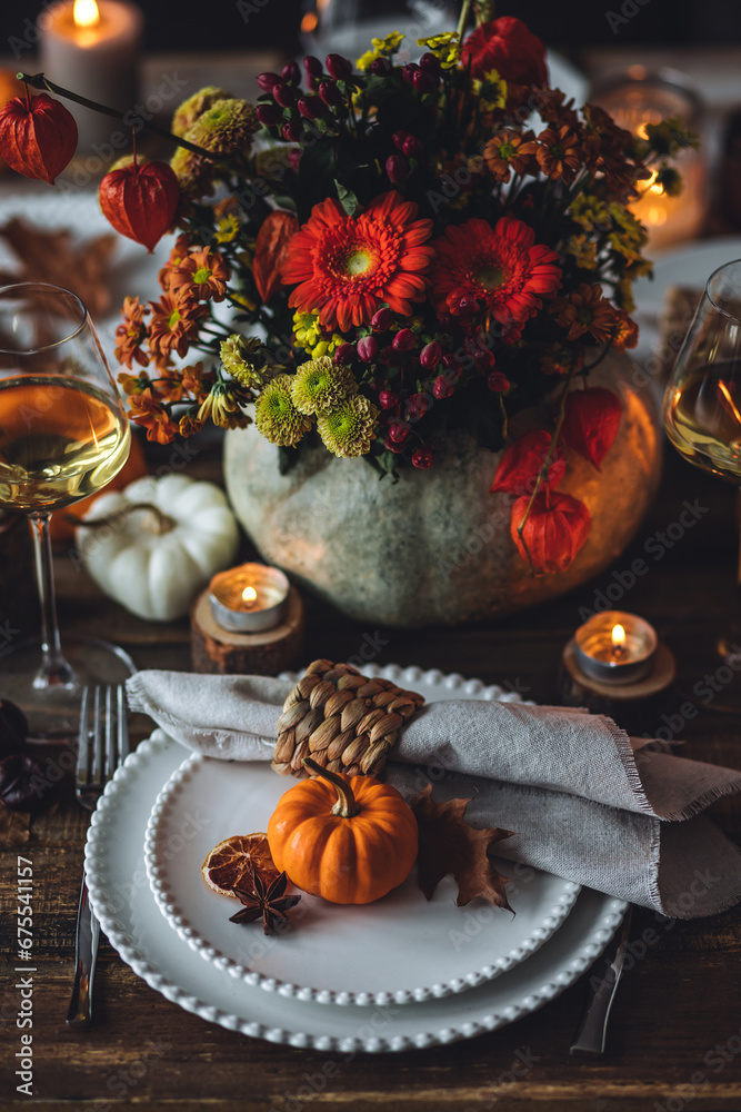 Elegant seasonal fall table setting with floral composition in a pumpkin vase as a centrepiece. White wine, burning candles, romantic cozy atmosphere of family dinner, private autumn wedding