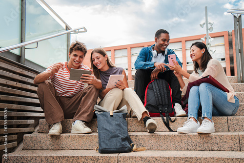 Group of multiracial high school students talking on a staircase at university campus, using a tablet app and smartphone to do homework project after class. Friendly teenage people studying together photo