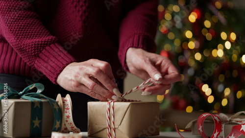 Obraz na plátně Woman in a burgundy sweater packs a lot of boxes with gifts in beautiful paper and ribbons, bows on the background of a Christmas tree
