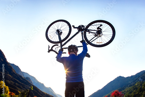 male cyclist with a raised bicycle against the backdrop of the sun and mountains
