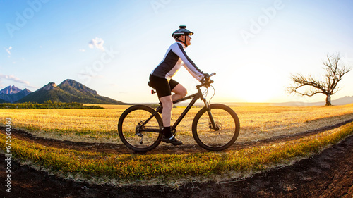 Fototapeta Naklejka Na Ścianę i Meble -  The cyclist rides a bike on the road near the field against the backdrop of the setting sun. Outdoor sports. Healthy lifestyle.