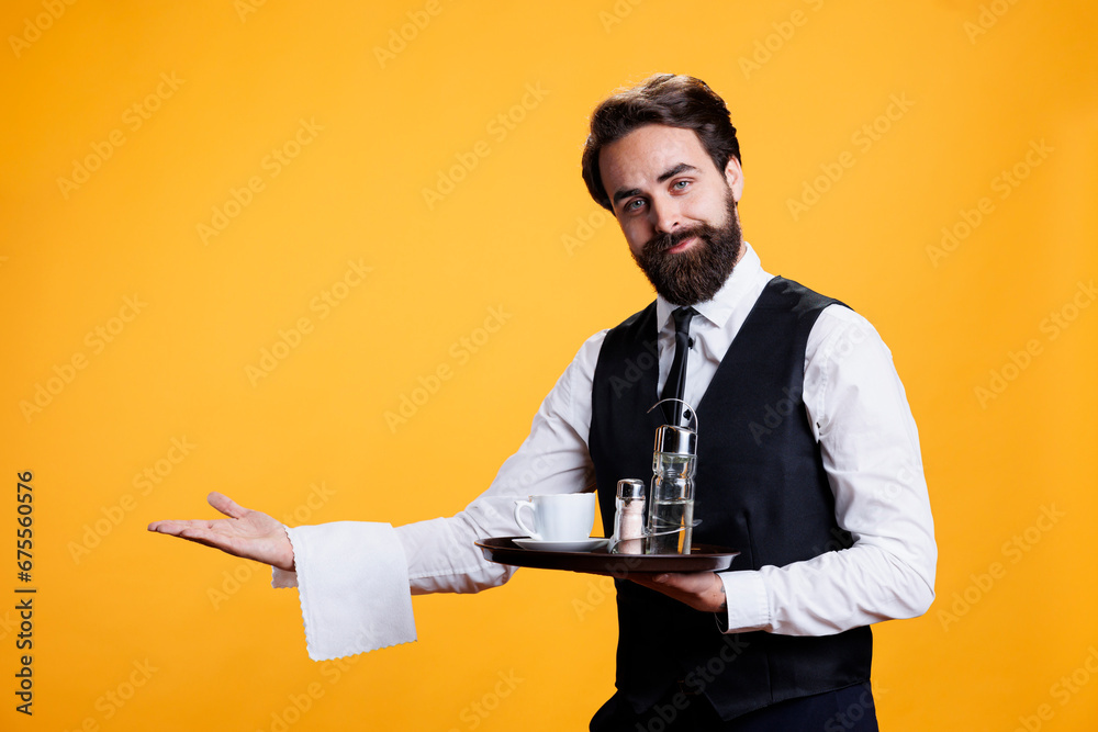 Skilled waiter shows direction to table at restaurant, serving people as occupation. Young man working as butler with meal tray in hand, pretends to serve cup of coffee to customers on camera. - obrazy, fototapety, plakaty 