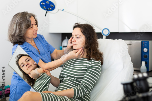 Experienced cosmetology doctor working in the clinic carefully examines the face of a young woman patient who is ..holding a mirror in her hands