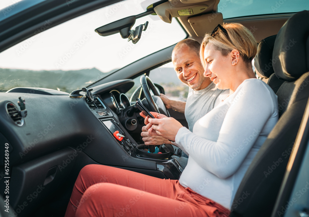 Happy smiling two young adulds couple using smartphone navigation inside car during auto jorney.They are laughing during road trip. Family, couples relations values, car sharing and traveling concepts Happy smiling two young adulds couple using smartphone navigation inside car during auto jorney.They are laughing during road trip. Family, couples relations values, car sharing and traveling concepts