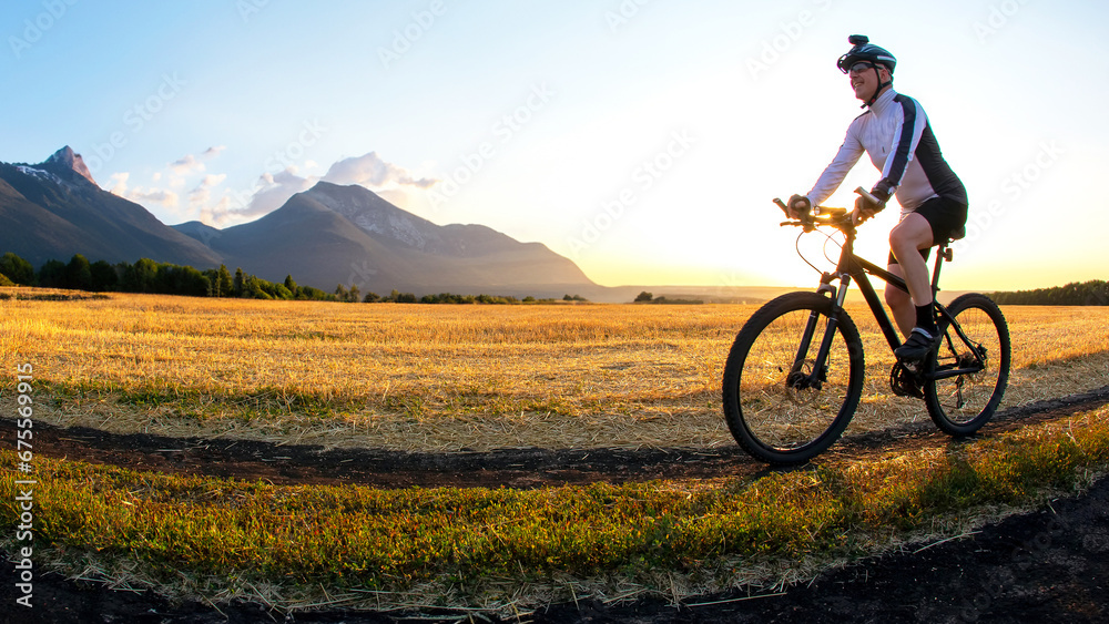 Fototapeta premium The cyclist rides a bike on the road near the field against the backdrop of the setting sun. Outdoor sports. Healthy lifestyle.