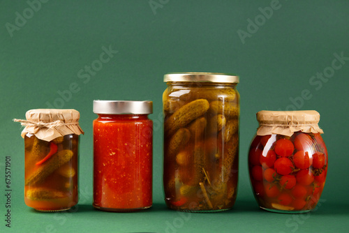 Canned vegetables in jars on a green background. Pickled cucumbers, tomatoes, chili sauce