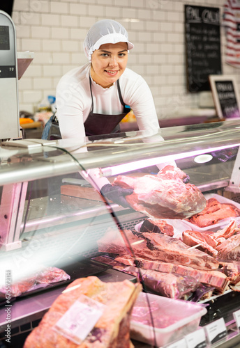 Young woman seller in uniform puts piece of beef in showcase