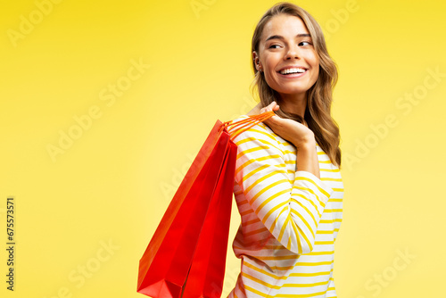Happy beautiful woman holding red shopping bags looking away isolated on yellow background, copy space. Shopping, sale, black Friday