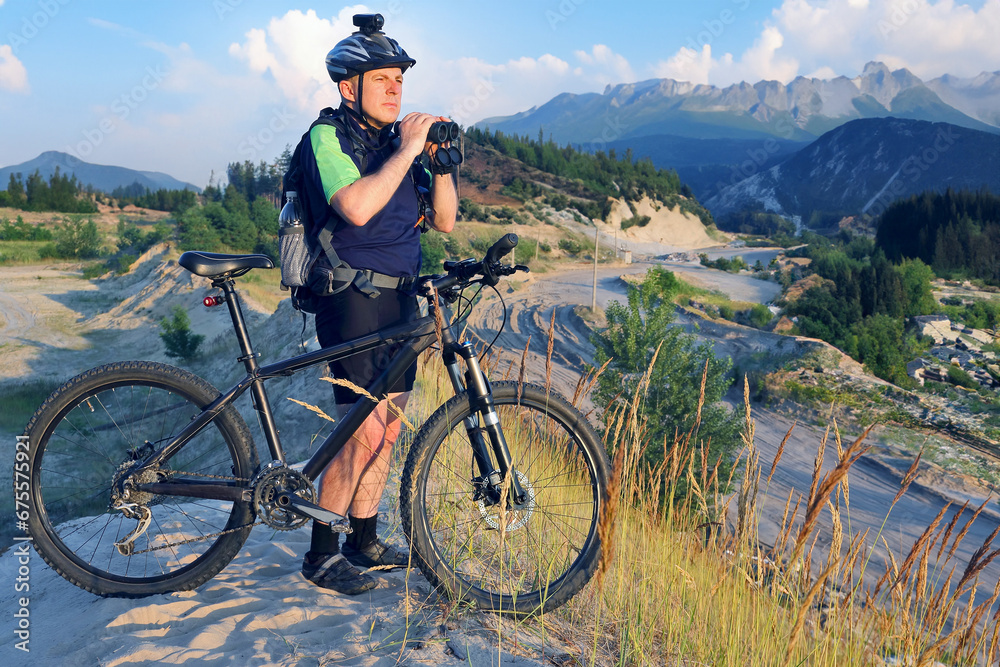 Fototapeta premium The cyclist with the Bicycle and binoculars in hand stands on a sandy hill in the sunlight.