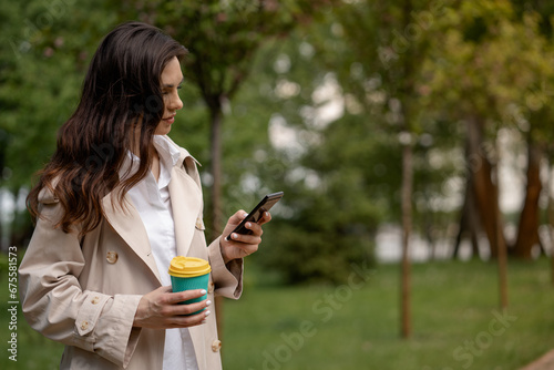 A young woman uses a smartphone, writes or reads a message, waiting to meet a friend or work colleague at the appointed time. A brunette lady walks in the spring park with a cup of coffee after