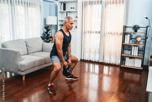 Athletic and sporty senior man engaging in leg day training session with squat and bodyweight kettle ball at home exercise as concept of healthy fit body lifestyle after retirement. Clout photo