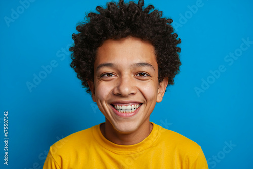 Cheerful young person with curly hair wearing a yellow shirt on a blue background, smiling broadly.