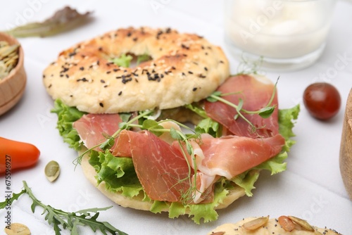 Tasty bagel with cured ham and lettuce on white tiled table, closeup