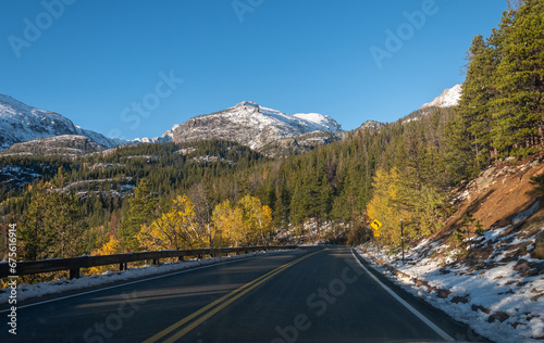 View looking out windshield of a car driving on a two lane mountain road in late autumn with golden yellow leaves and pine trees and a clear blue sky. Snowy mountains rise in the distance.