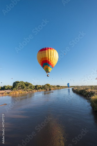A vibrant multicolor hot air balloon flies low over the Rio Grande River, its reflection visible in the surface of the water. More balloons can be seen further upriver in the distance.
