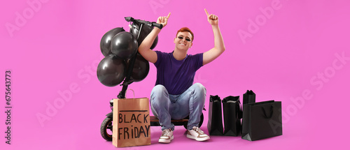Happy young man with kick scooter, balloons and shopping bags pointing at something on magenta background. Black Friday sale