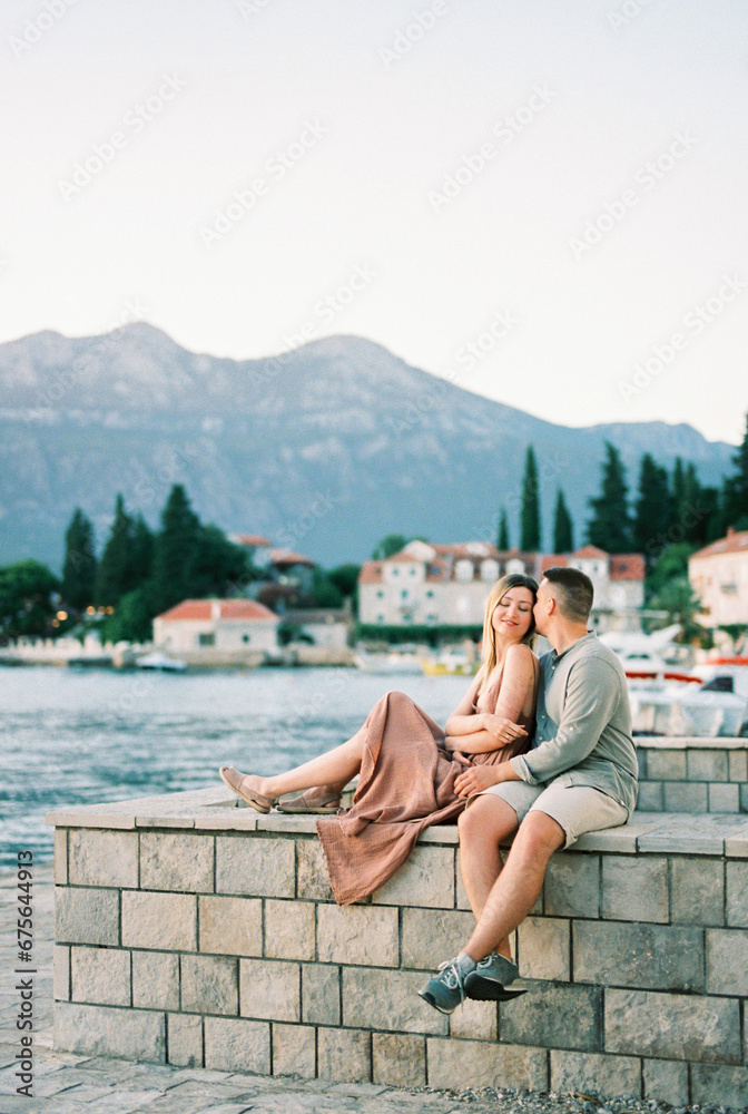 Man kisses woman on the temple while sitting with her on a stone fence on a pier by the sea
