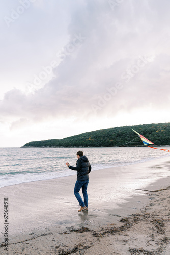 Man with a colorful kite on a rope walks along the sandy beach towards the sea. Side view