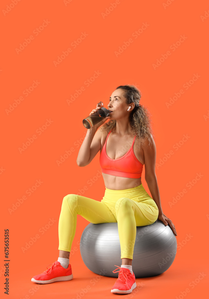 Sporty adult woman drinking water from bottle while sitting on fitness ball against orange background