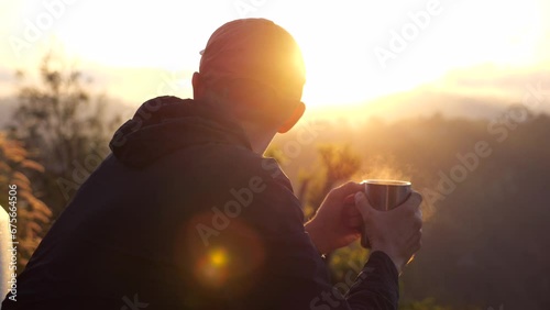 Hiker man sitting on chair, holding coffee mug with steam at dawn. Back view of young traveler that woke up early morning to meet dawn and bathe in sunshine. Concep travel on camping van to meet dawn.