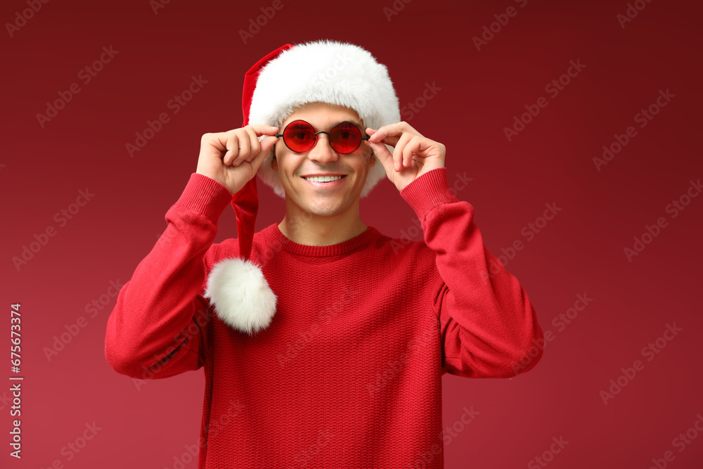 Young man in Santa hat with glasses on red background Young man in Santa hat with glasses on red background