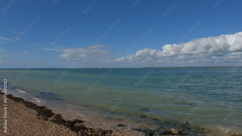 Cristal clear water waves gently kissing the orange pebble beach in the Isle of Wight. Steady camera with the blue sky and some white fluffy clouds in the background. Cristal clear water waves gently kissing the orange pebble beach in the Isle of Wight. Steady camera with the blue sky and some white fluffy clouds in the background.