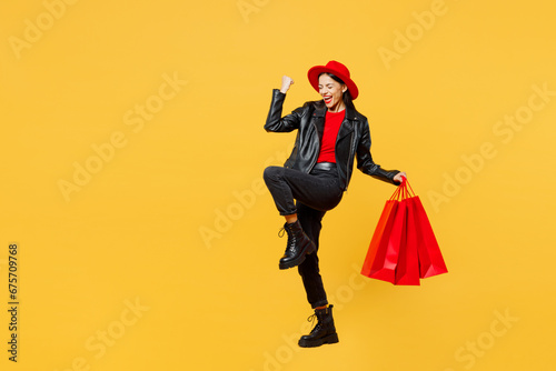 Full body side view young woman wear casual clothes red hat hold shopping package bags do winner gesture celebrate clench fists isolated on plain yellow background. Black Friday sale buy day concept.