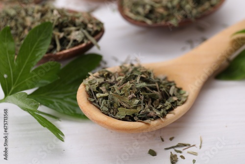 Spoons with dried aromatic parsley and fresh leaves on white wooden table, closeup